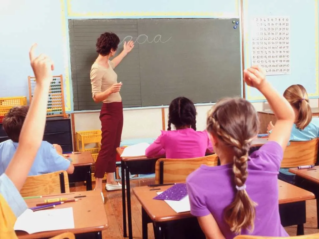 Profesora enseñando en clase mientras estudiantes levantan la mano, ilustrando objetivos didácticos en acción.
